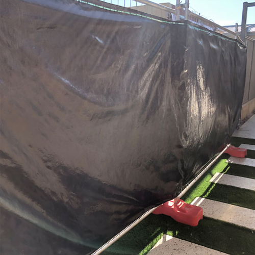 Temporary fencing covered with a black tarp-like material, secured in place with weighted red fence feet, situated on a construction site.